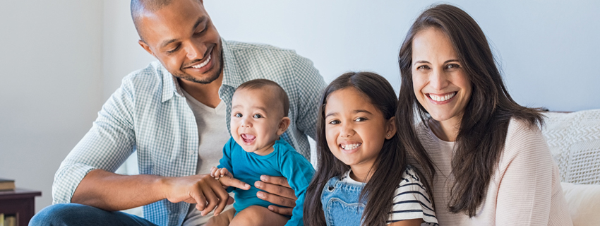 Young family smiles at camera. Dad is holding baby and mom sits with older daughter.