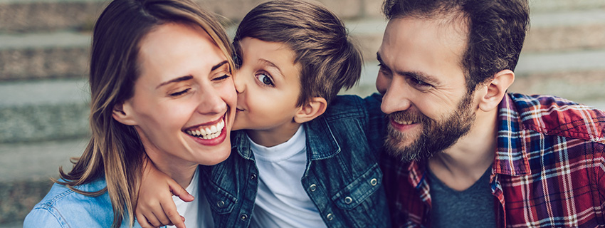 Boy hugging mom and dad while giving mom kiss on cheek.