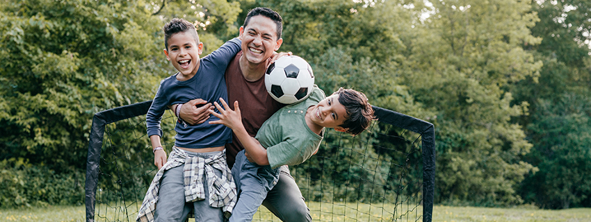 Dad hugging two sons in front of soccer goal. One of the boys is holding a soccer ball.
