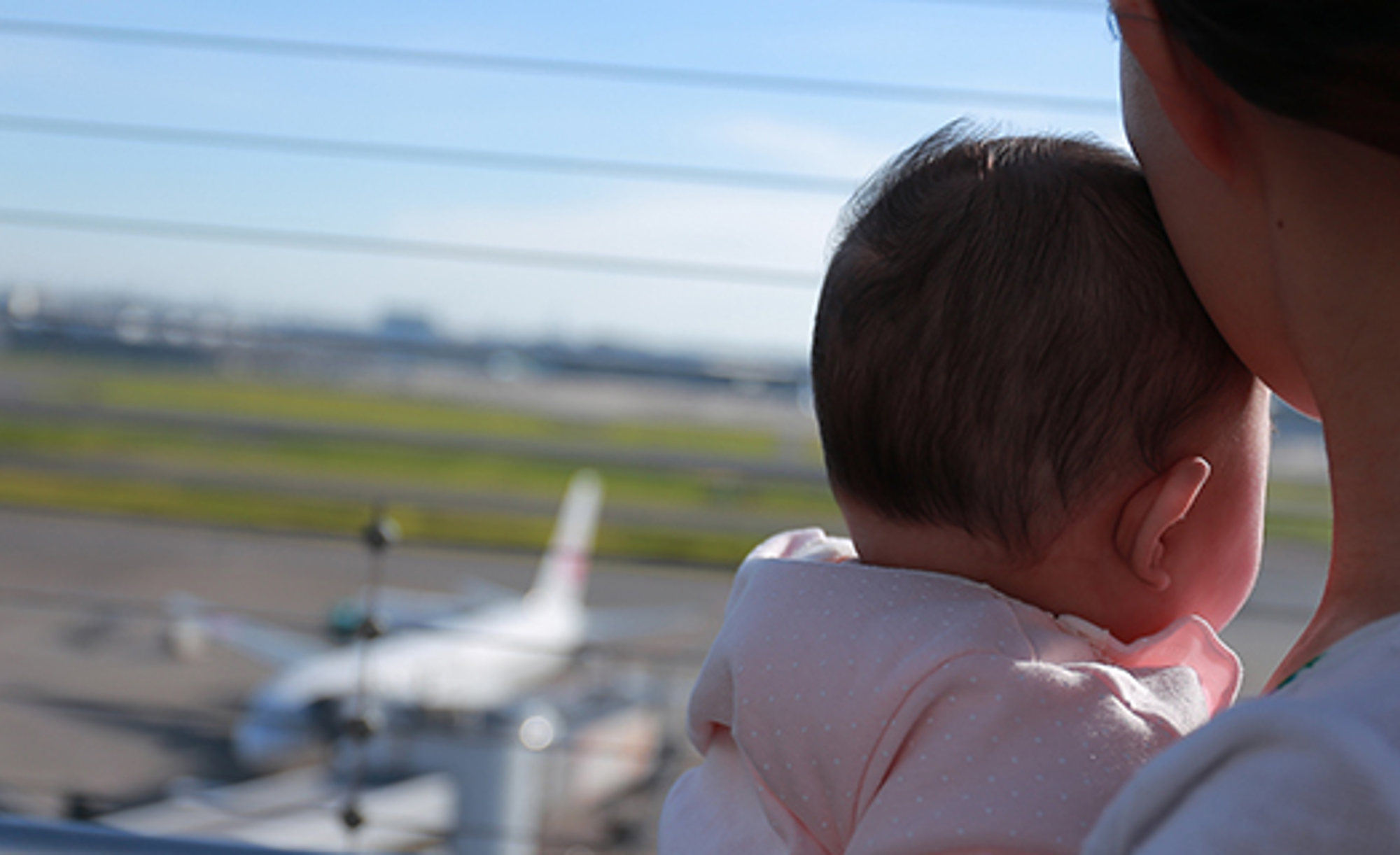 Mom holds baby while looking out window at airplane. 